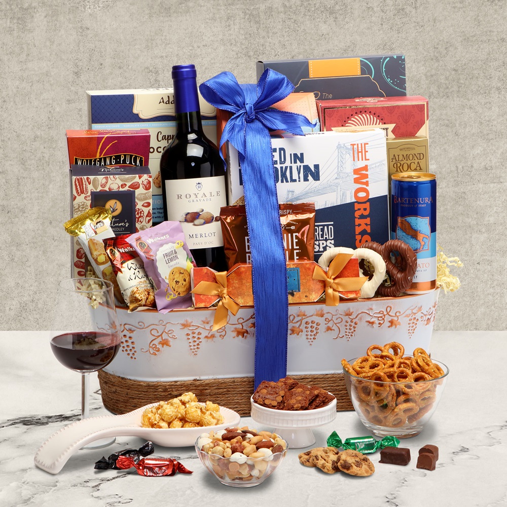 A bright blue ribbon ties together a wicker and metal basket combo filled with seasonal holiday treats, next to a glass of red wine and plates full of popcorn, brittle and chocolate